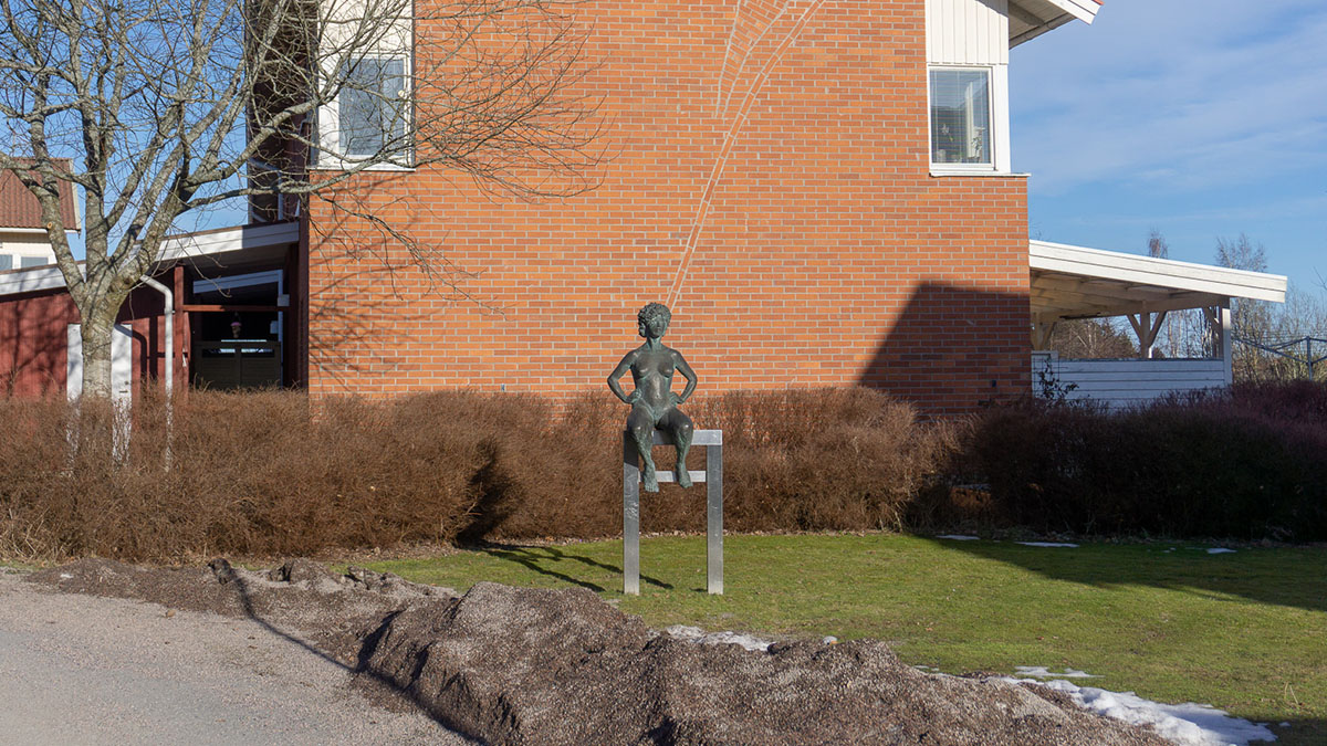 Staty av sittande kvinna, framför tegelvägg.
