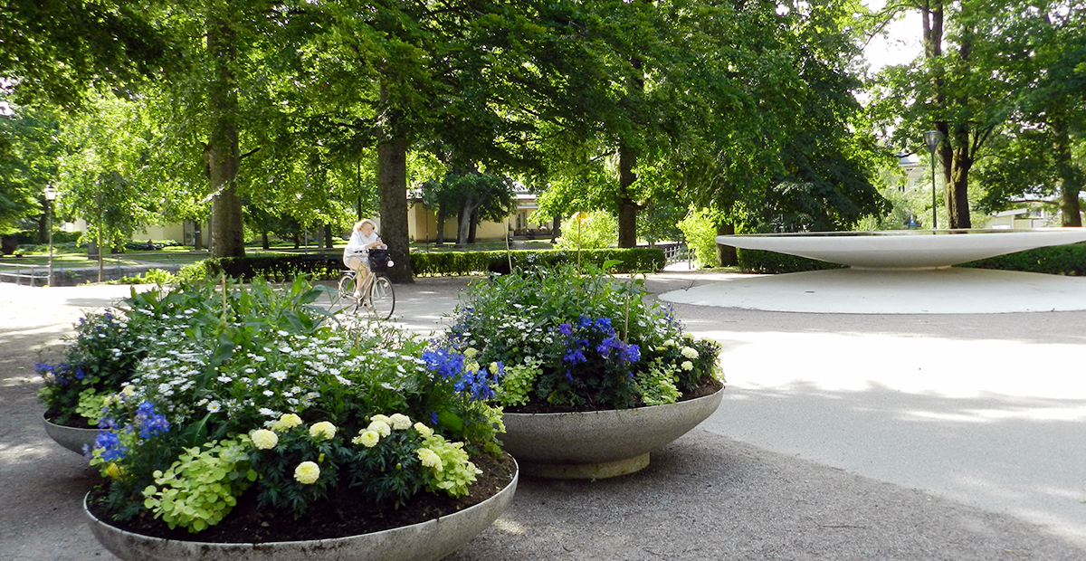 Del av Slottsparken med cyklist och tre skålformade planteringskärl i betong, med blommor i lime, vitt, blått och gult. Kärlen är cirka 1 till 2 meter i diameter. 